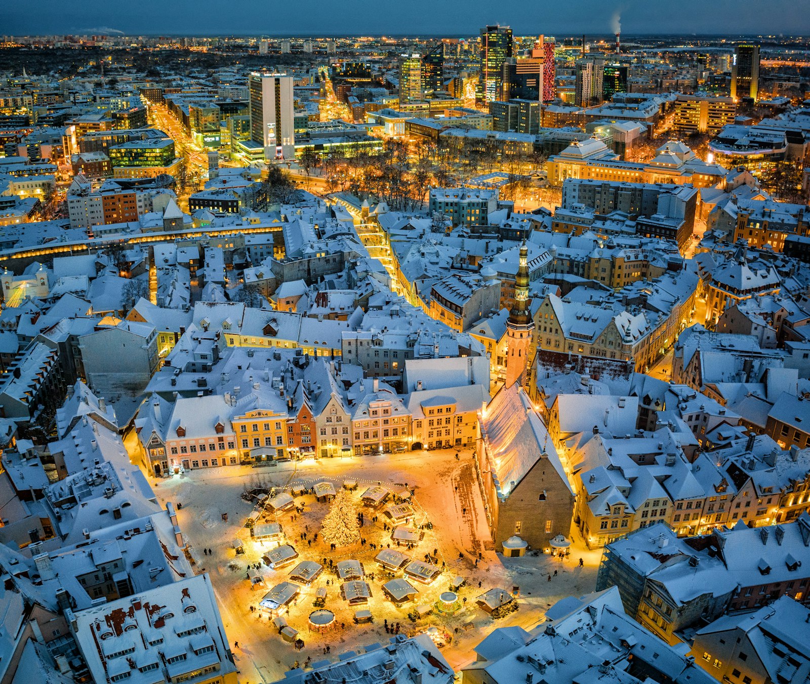 La vieille ville de Tallinn sous la neige, la nuit, la place médiévale éclairée par son marché de Noël, avec le quartier d’affaires moderne derrière.